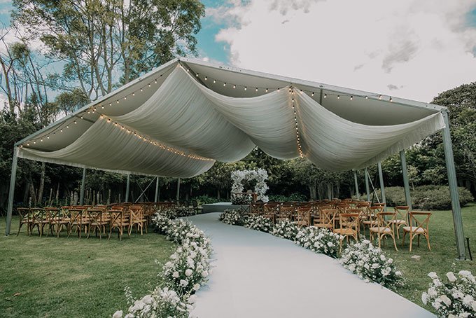 Elegante montaje para ceremonia de matrimonio campestre, con un pasillo blanco al aire libre bajo una carpa con telas y luces cálidas.