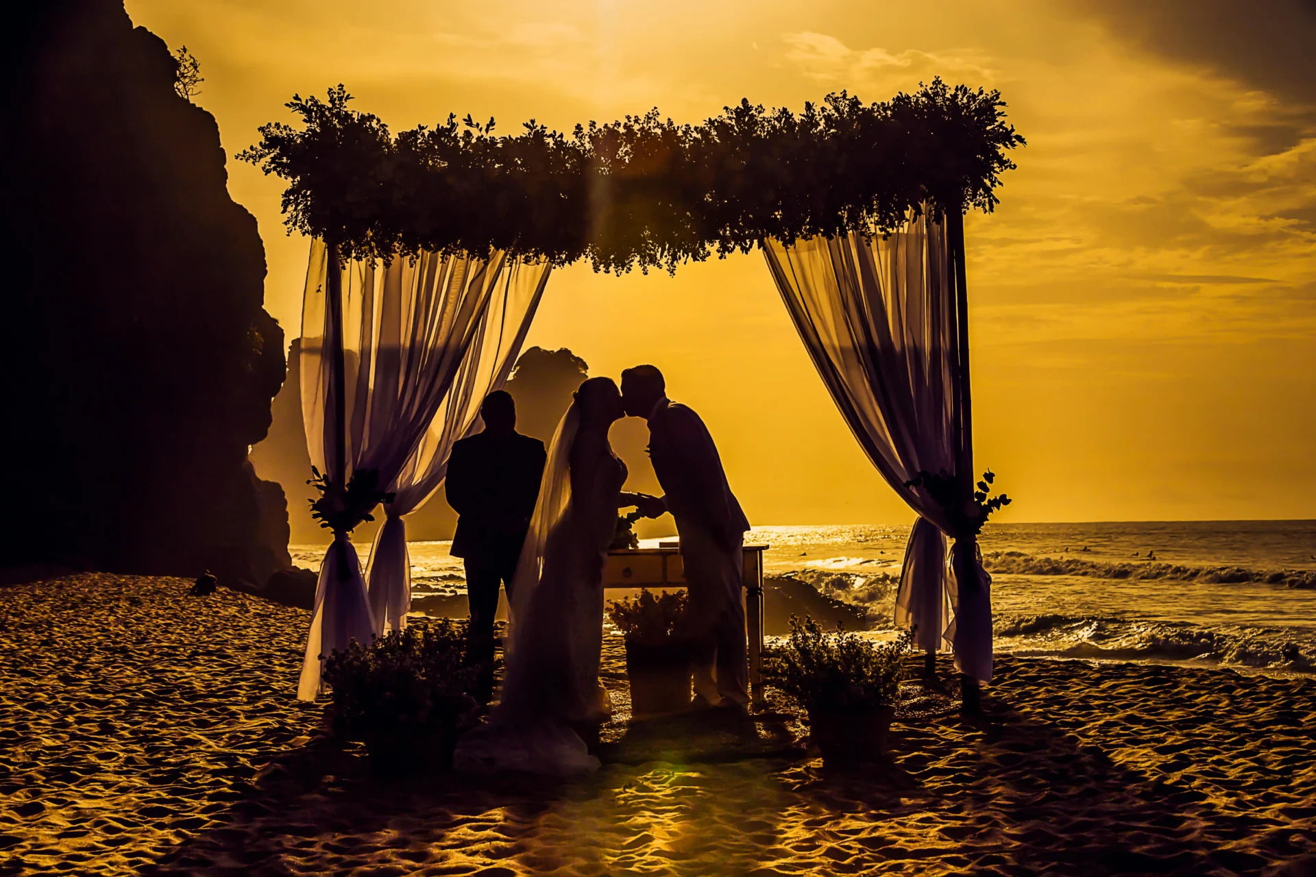 Silueta de pareja en boda espiritual en la playa al atardecer, besándose bajo un altar con telas en Colombia