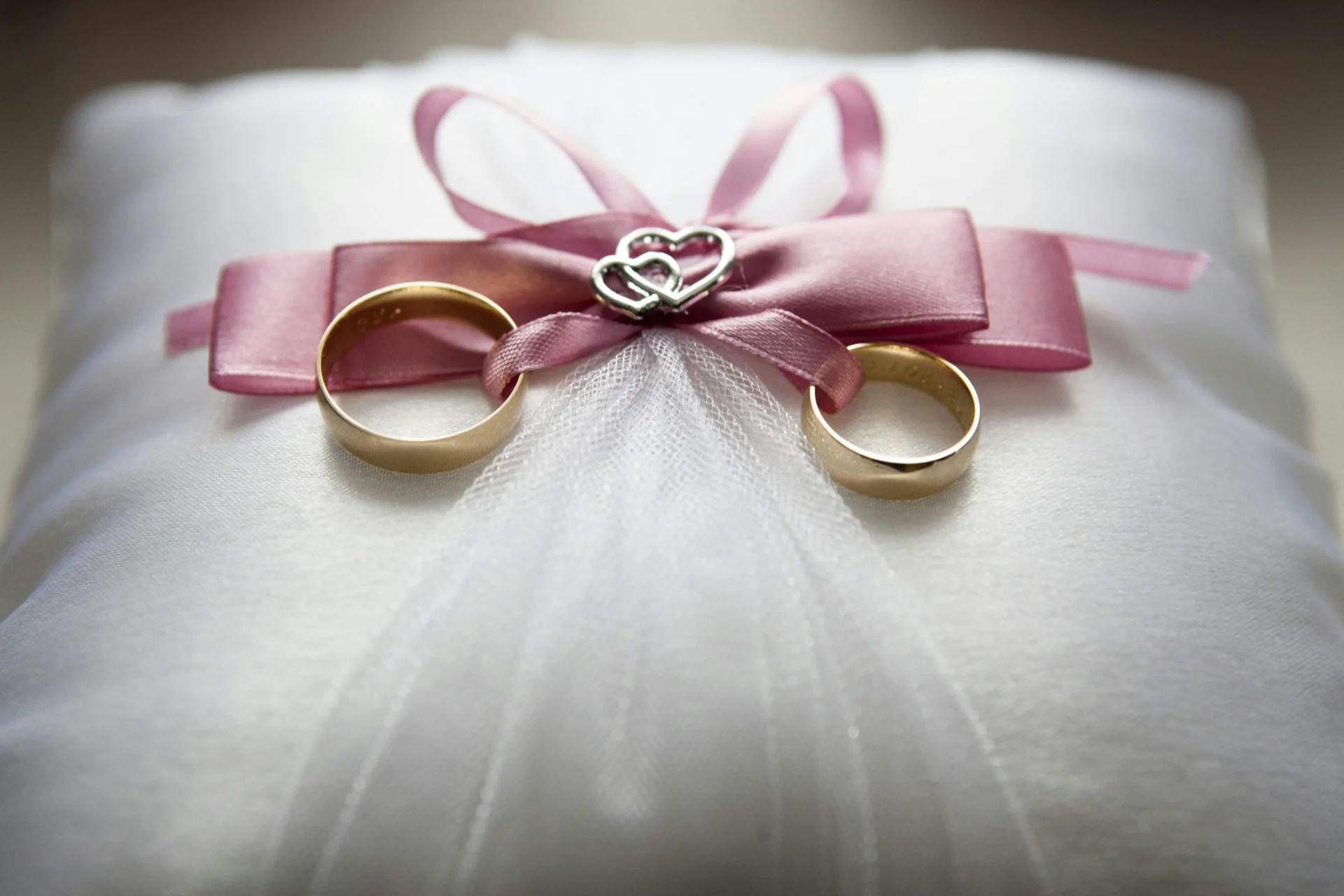 dos anillos de boda dorados sobre un velo blanco, adornados con una cinta rosa y un dije de corazón, simbolizando un aniversario de lino
