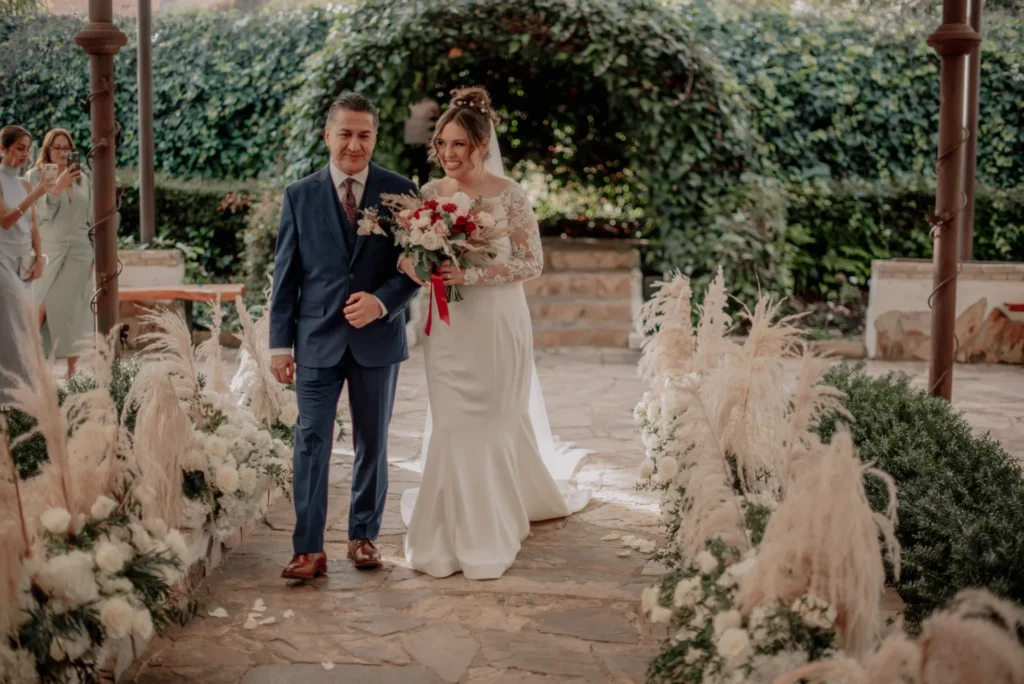 Novia sonriente caminando del brazo de su padre por un pasillo de piedra decorado con flores y hierba de pampa, durante su ceremonia de boda al aire libre en Zipaquirá.