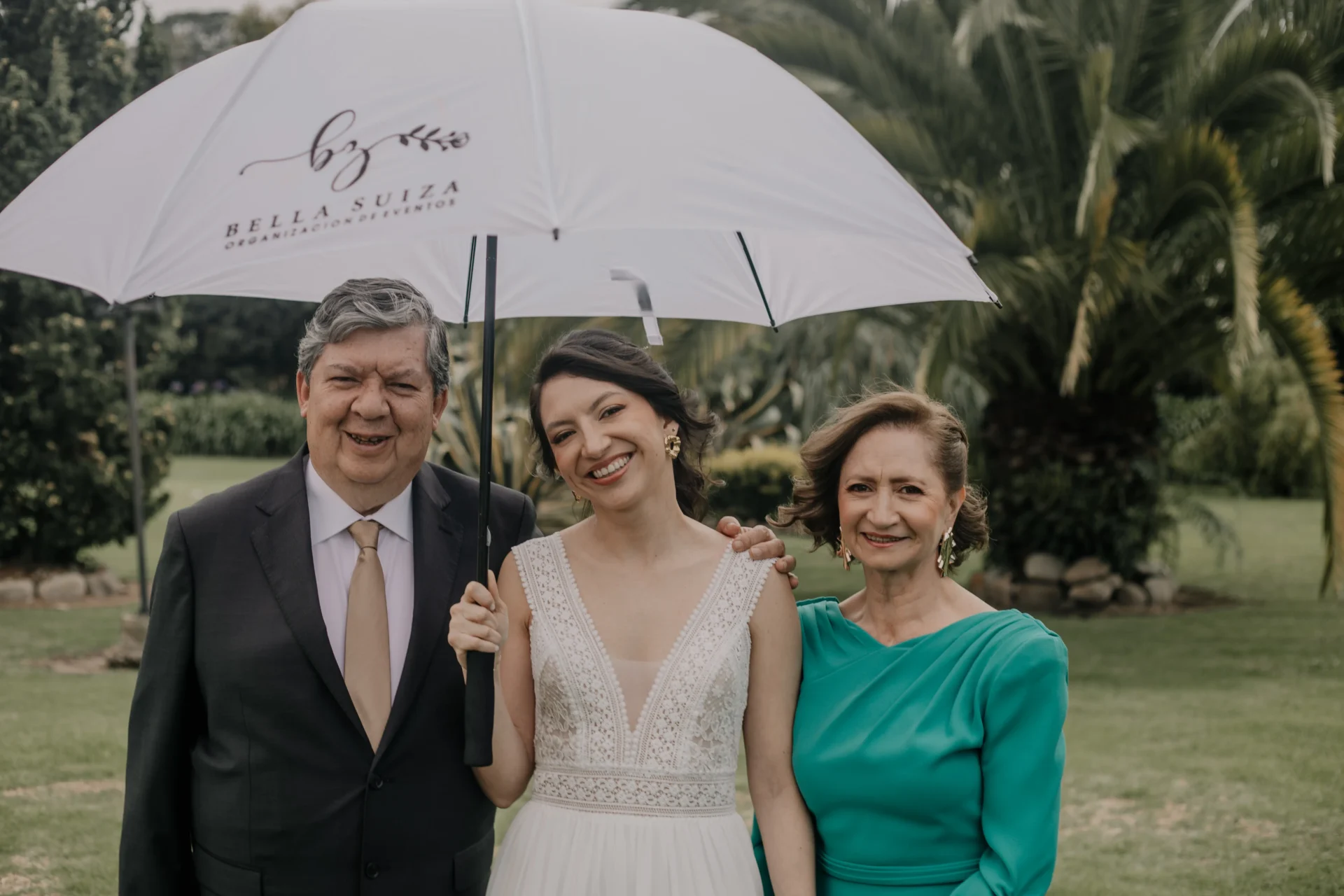 Novia juntos a sus padres con una sombrilla protegiéndose del sol pronta para presentase al altar.