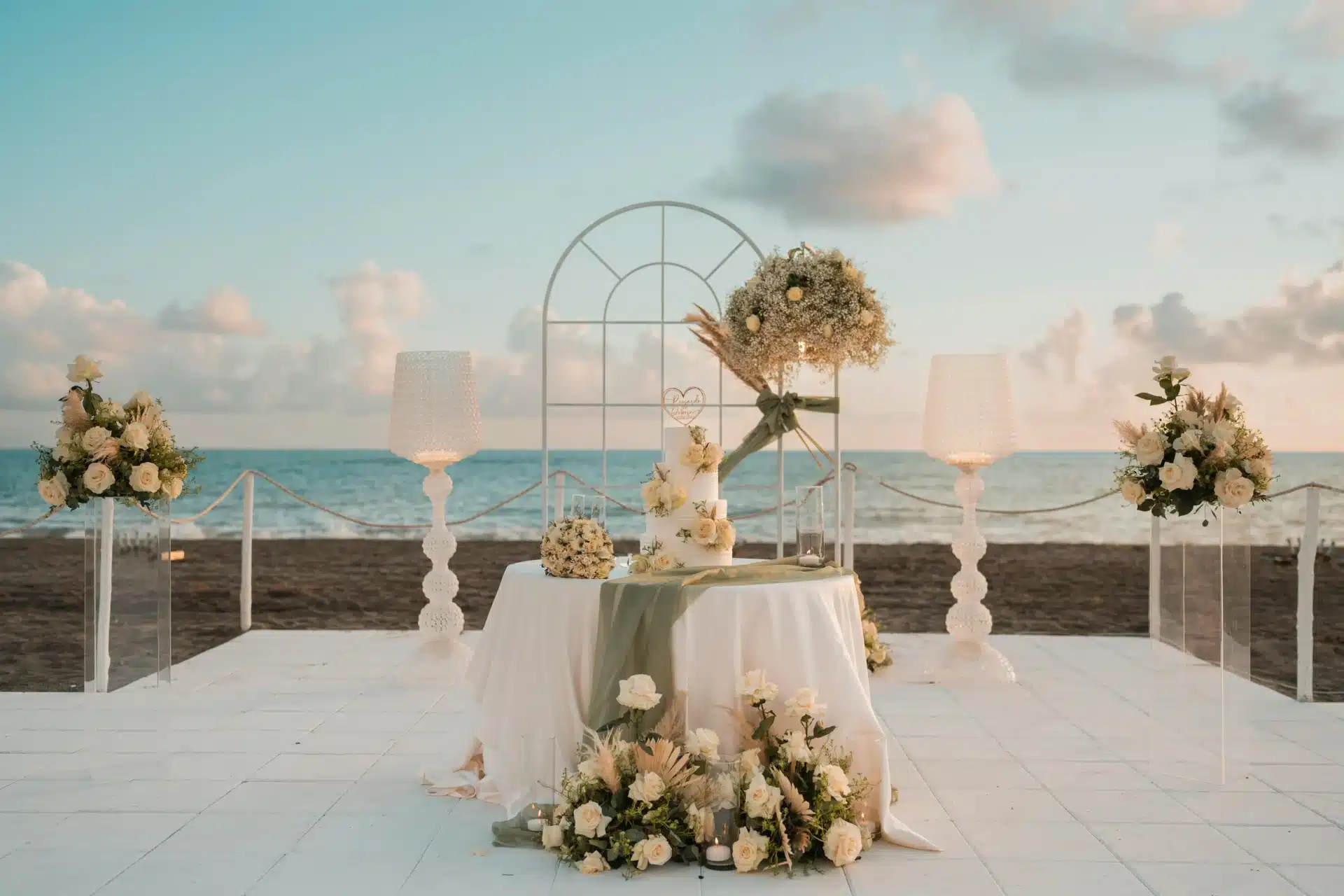 : Arreglo de boda elegante frente al mar en San Andrés, con mesa decorada, flores blancas y arco minimalista sobre una plataforma al aire libre, con el océano y cielo al fondo.