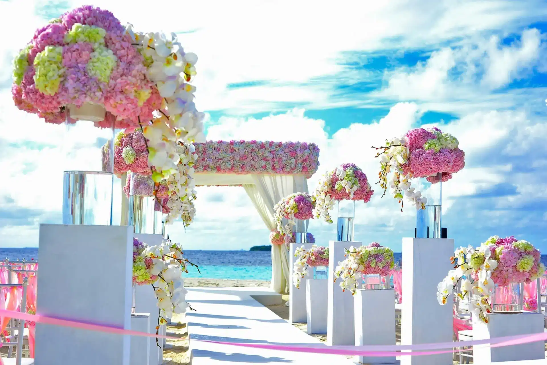 Ceremonia de boda al aire libre en la playa de San Andrés, con pasillo decorado con flores rosas y blancas, columnas florales y arco nupcial frente al mar Caribe bajo un cielo azul.